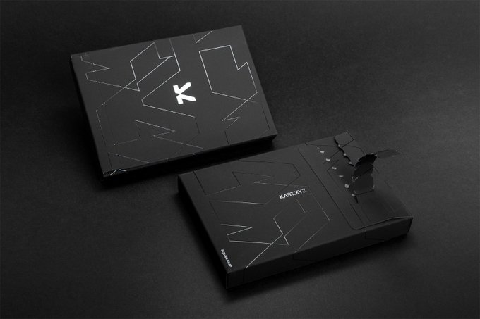 Two sleek black boxes featuring geometric line designs and the "KARATXYZ" branding sit on a dark surface, with the top box displaying a stylized "K" logo, complementing the announcement of physical cards being shipped to members who reached a new tier.