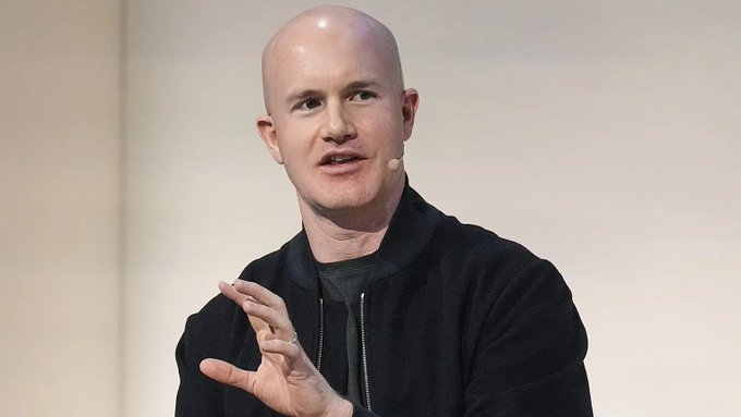 A bald man wearing a black jacket and headset microphone gestures with his hand while speaking, identified by the accompanying article as Brian Armstrong, CEO of Coinbase, who is discussing regulatory clarity for crypto and calling on the SEC and CFTC to take action after the failure of the CLARITY Act.