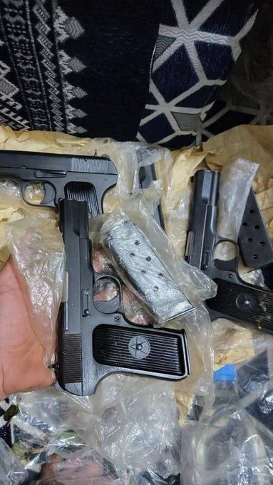 A hand holds two black handguns and several loaded magazines wrapped in plastic bags against a patterned fabric background.