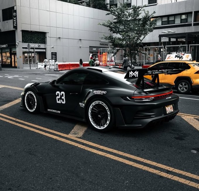 A matte black Porsche 911 GT3RS with the number 23, Michelin branding, and a large rear wing is parked on a city street, with a yellow NYC taxi visible in the background.