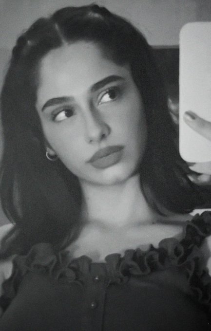 A black-and-white selfie features a woman with dark hair and hoop earrings taking a mirror photo with a white phone.