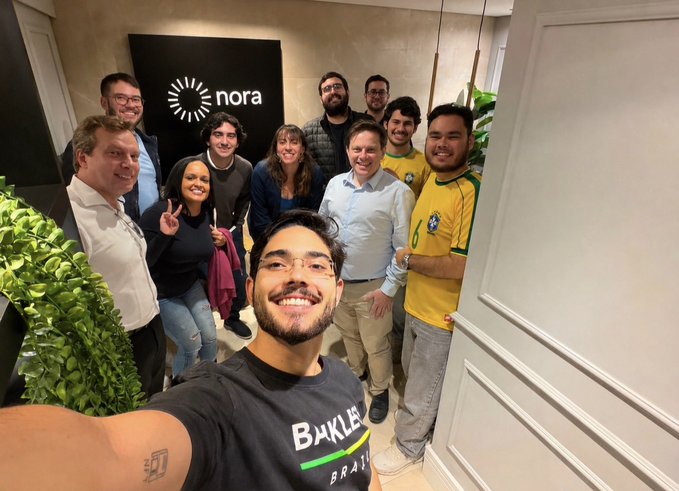 A selfie of a team smiling in an office with a "nora" logo on the wall behind them, one person wearing a Brazil national team jersey. The caption highlights a visit to the nora finance team in São Paulo and congratulates them on their honorable mention by Colosseum.
