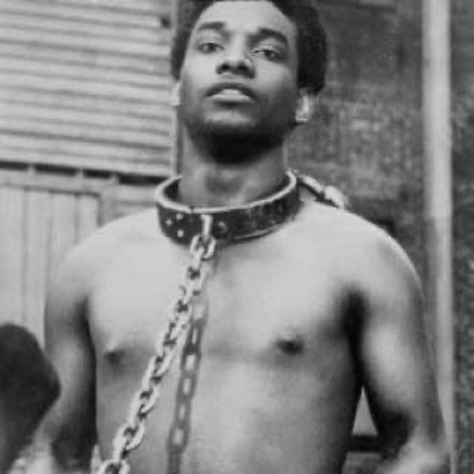 A black-and-white photo shows a shirtless man wearing a chain and collar, with a stoic expression, standing outdoors in front of a corrugated metal wall. The image appears to be a still from a film or TV scene and is being used to humorously illustrate the post's joke about someone overworking for the Solana Foundation.