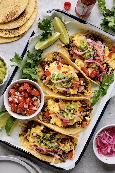 A tray of hard-shell tacos topped with cheese, guacamole, salsa, and pickled red onions, served with lime wedges, pico de gallo, guacamole, and extra tortillas on the side.
