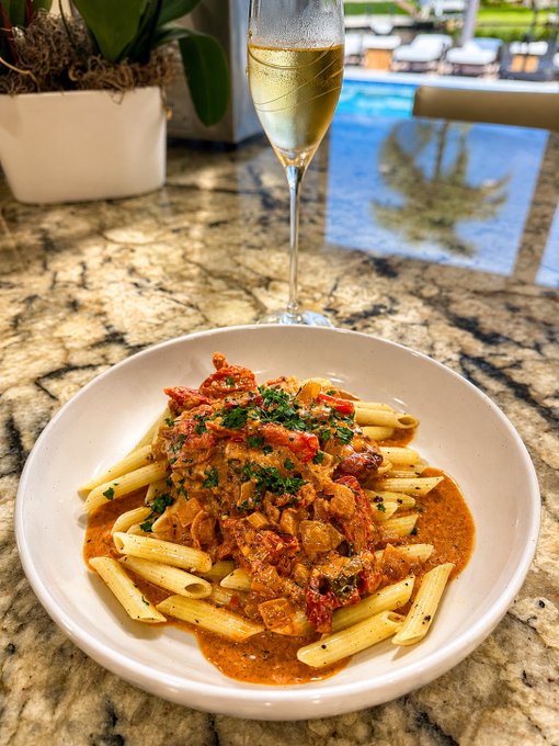 Penne pasta topped with creamy tomato-sauced Marry Me Chicken, garnished with fresh parsley, is served poolside alongside a glass of sparkling wine.