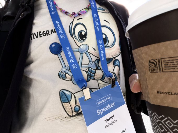 A person wears a t-shirt featuring a cartoon robot mascot alongside a "Speaker" badge for Yohei Nakajima at a Runlayer/World's Fair event, while holding a coffee cup. The shirt's "TIVEGRAPH" text appears to be cut off but likely reads "ACTIVEGRAPH," matching the "activegraph" branding mentioned in the post.