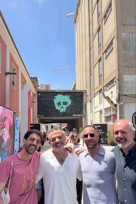 The image features a group of four men standing in front of an open door, possibly an art gallery or building, as indicated by the "Kodafone" logo displayed on one of them. The man with a camera around his neck can be seen capturing the moment, and he is likely the first to arrive at the location. 

The other three men are standing close together, each in their own stance but all facing towards the same direction or towards an interesting object that might be behind the door. A handbag is visible near one of them, possibly belonging to someone who has just arrived or is preparing to leave.