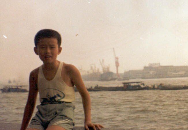A young boy poses by the waterfront in Hong Kong, with cargo ships and cranes visible across the harbor. The post suggests this is a childhood photo of the man who later became the founder of the world's largest Bitcoin exchange.