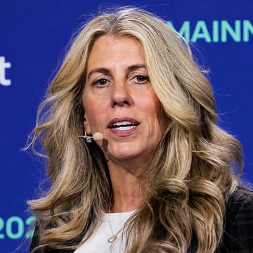 A close-up of a blonde woman wearing a headset microphone speaking at an event with a blue background partially showing the word 'MAINE'.