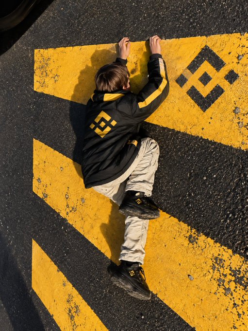 A person in a black and yellow Binance hoodie and matching sneakers lies on a road marked with yellow Binance logos, visually reinforcing the message "I'm literally sticking with Binance." The branded pavement and apparel symbolize unwavering loyalty to the cryptocurrency platform.