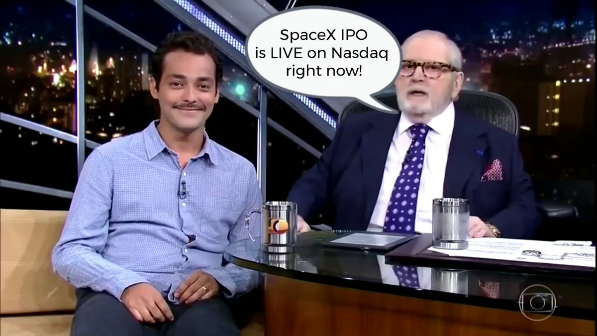 The image shows two men sitting at a desk, with one man wearing glasses and the other wearing a suit. They are both smiling as they appear to be discussing something related to the SpaceX IPO. The background of the image features a city skyline, indicating that this conversation might have taken place in an urban setting or during a special event like the launch of a new space project.