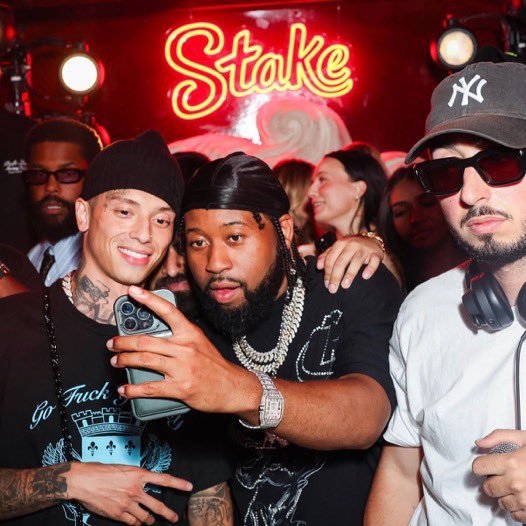 Two men take a group selfie at a lively club under a glowing red "Stake" neon sign, with one holding up a smartphone to capture the moment while wearing a Yankees cap and dark sunglasses. The vibrant nightlife scene, filled with stylish patrons and bold branding, ties into the post's playful reference to a "Stake Finale" that outshines major sporting events.