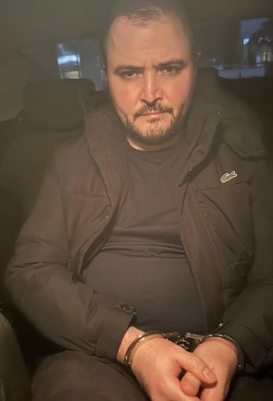 A man with a beard wearing a dark puffer jacket sits in handcuffs inside what appears to be a vehicle.
