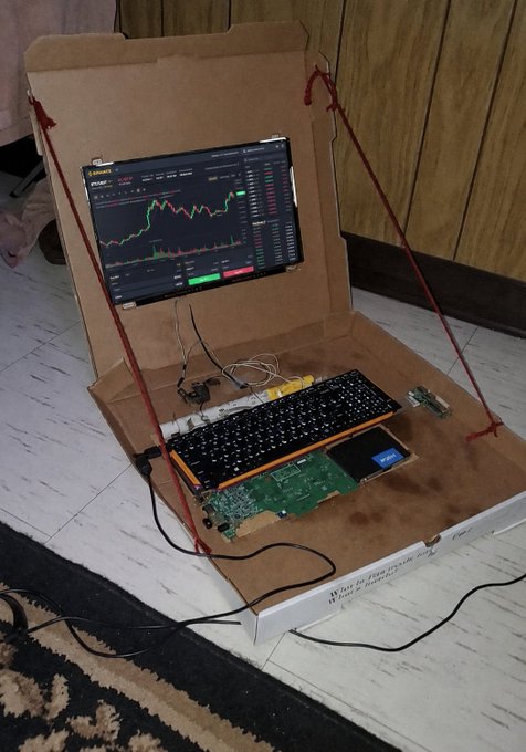 A makeshift DIY trading station built from a cardboard box displays a monitor showing a Binance trading chart, with the post's Binance referral URL ("accounts.binance.com/en/register") promoting crypto trading sign-ups. The image reflects the "simple possibilities" of trading from anywhere with basic materials, paired with ambitious financial goals in the crypto market.
