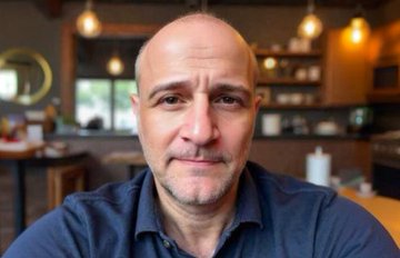 A selfie of a bald man in a blue shirt sitting in what appears to be a cafe or restaurant with pendant lights and wooden furniture in the background.