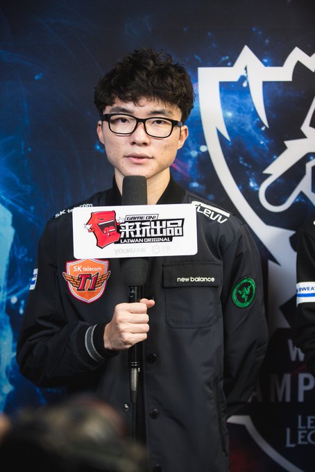 A man with curly hair and glasses being interviewed, wearing a black jacket with SK telecom T1 and League of Legends logos while holding a microphone labeled 'GAME FPV'.