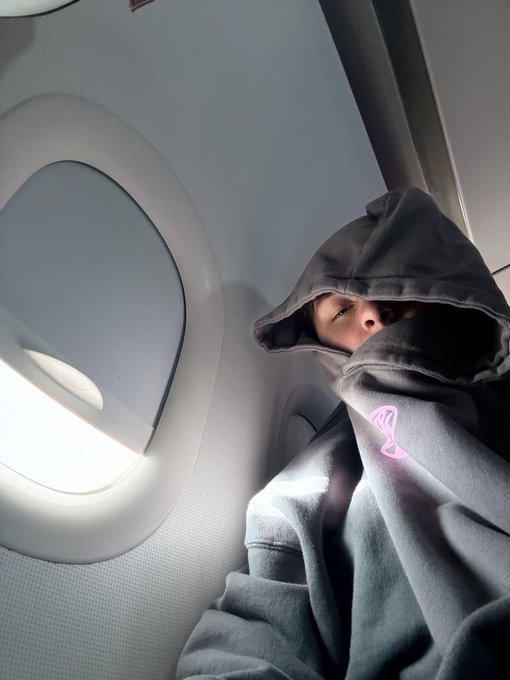 A person bundled in a grey hoodie with a pink logo sits by an airplane window.