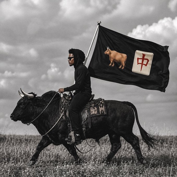 A person in all black rides a large black bull across a grassy field, holding a black flag featuring a golden bull and a Mahjong tile with the red Chinese character "中" (meaning "middle" or "red dragon"). The image visually echoes the post's "bullish" sentiment, using the rising bull, determined rider, and rising flag as a metaphor for the optimistic outlook on holding $ANSEM.