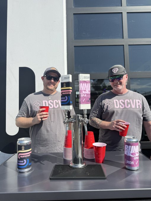 Two men stand behind a table with beer taps, wearing matching gray 'DSCVR' branded t-shirts and holding red plastic cups, with cans of 'Bluetopia' beer displayed.