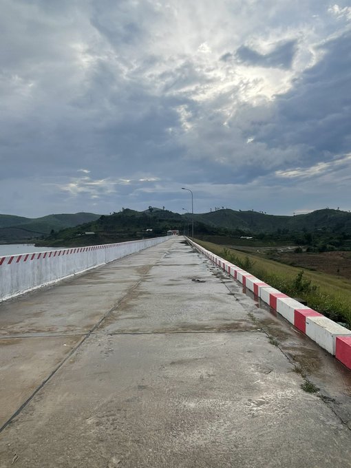 A scenic view of an empty concrete road or bridge stretching into the distance, bordered by red and white painted barriers, with rolling green hills and a cloudy sky in the background.