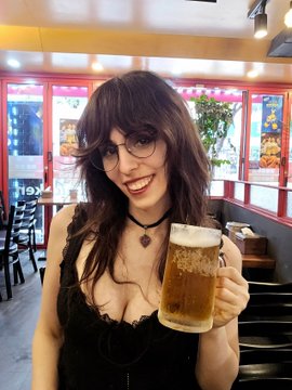A smiling woman with dark hair, glasses, and a black choker holds up a large mug of beer in what appears to be a restaurant.