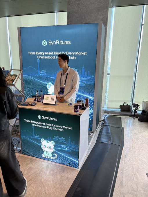 A booth for 'SynFutures' displaying the slogan 'Trade Every Asset. Build for Every Market. One Protocol. Fully Onchain.', staffed by a smiling man in a striped shirt behind a counter with QR codes and merchandise.