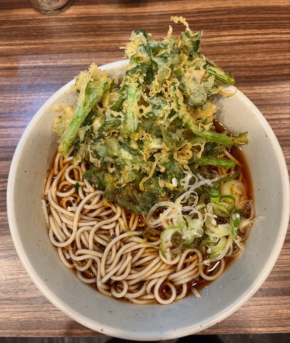 A bowl of soba or ramen noodles in dark broth, topped with a generous portion of crispy kakiage (tempura fritters) containing green vegetables, served with sliced green onions.