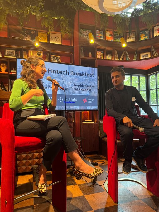 A woman speaks into a microphone while seated across from a male guest on red chairs during the "Fintech Breakfast" panel at a Midnight Foundation event, with on-screen text identifying her as Sophia Furber, S&P Global. The setting appears to be an intimate, warmly lit venue with bookshelves and hanging plants, aligning with the post's reference to "Midnight Monument Bank" as the hosting organization.