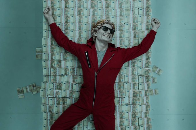 A person in a red jumpsuit and sunglasses poses with arms raised in front of a wall completely covered in stacks of cash.