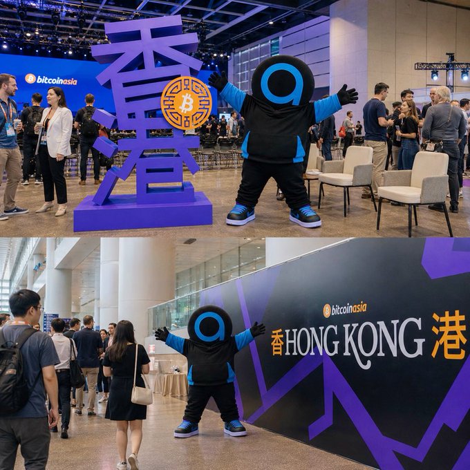 The mascot, dressed in a black and blue outfit with a distinctive circular logo, poses in front of a large purple "bitcoin" sign and a "HONG KONG" wall featuring the @bitcoinasia branding, capturing the lively atmosphere of the Bitcoin Asia conference referenced in the caption.