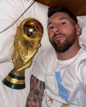 A selfie of soccer player Lionel Messi lying in bed next to the FIFA World Cup trophy, wearing an Argentina national team jersey.