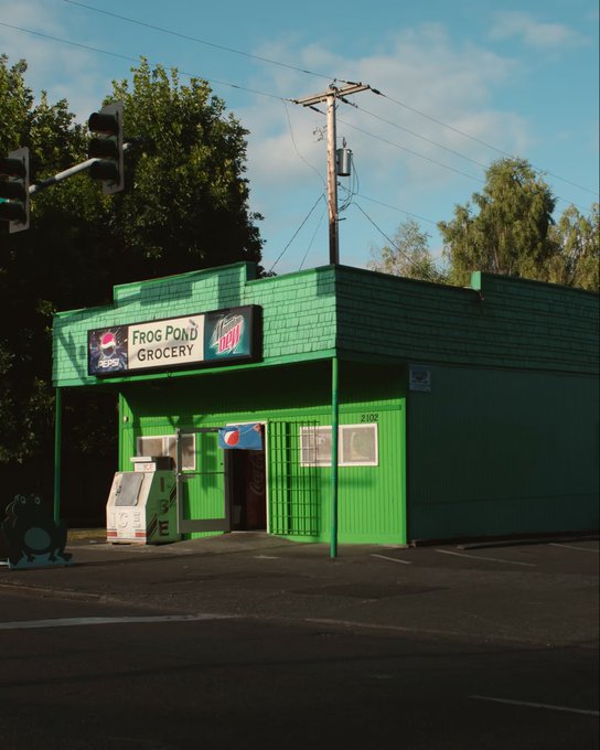 A green-painted corner store named 'Frog Pond Grocery' with a '7up' and 'Mountain Dew' sign, featuring a Pepsi vending machine outside and a frog statue on the sidewalk.
