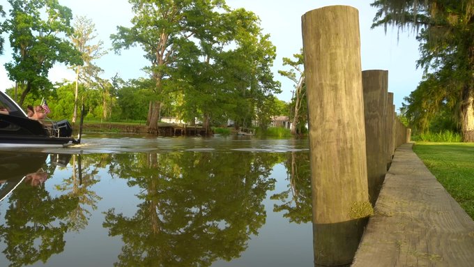 A scenic view of a calm river lined with wooden pilings, with trees draped in Spanish moss and a boat visible on the left, possibly along a bayou.