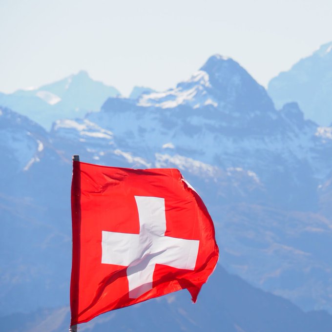 The Swiss flag waves in front of snow-capped Alpine peaks, symbolizing the Swiss central bank's behind-the-scenes accumulation of over 760,000 MicroStrategy shares—worth roughly $140 million—as a quiet, indirect route into Bitcoin exposure since 2024.