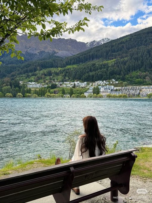 A woman with long dark hair sits on a wooden bench looking out over a turquoise lake toward a town nestled at the base of forested mountains.