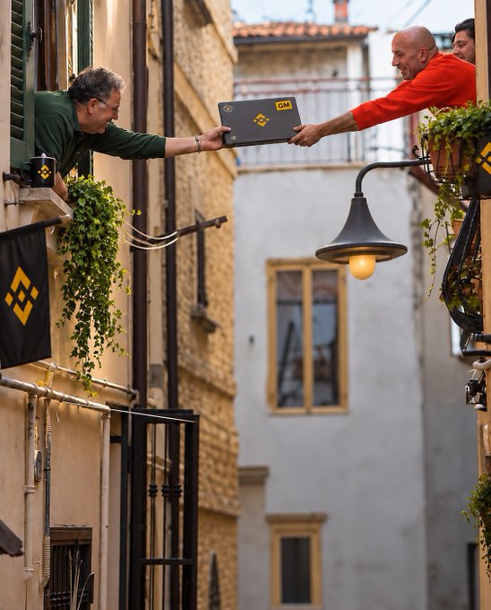 Two men lean out from opposite balconies across a narrow alleyway, passing a black folder marked "GM" in bold yellow letters between them. The image humorously captures the "brb" moment of someone stepping away to hand off market-watching duties to a neighbor, suggesting a casual, neighborly exchange of responsibility.