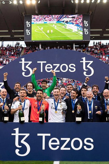 Real Madrid football players celebrating a championship victory with trophies, standing in front of a Tezos sponsor banner on what appears to be a podium stage.