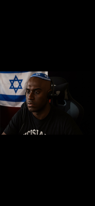 A man wearing a blue kippah and headphones sits in front of an Israeli flag.