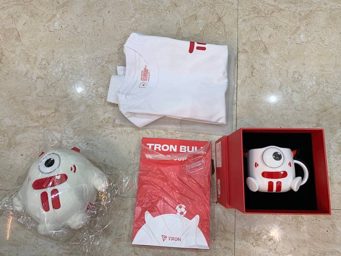 A collection of 'TRON BURGER' merchandise laid out on a tiled floor, including a folded white t-shirt, a plush toy of a white one-eyed character, a red booklet, and a matching character mug in a red presentation box.