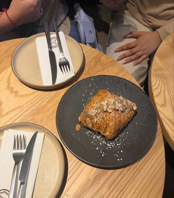 A pastry, appearing to be an almond croissant, sits on a black plate dusted with powdered sugar, alongside two place settings with cutlery on a wooden table.