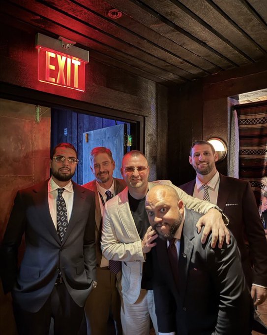 A group of five men in suits pose together near an illuminated red "EXIT" sign above a doorway in a dimly lit venue.
