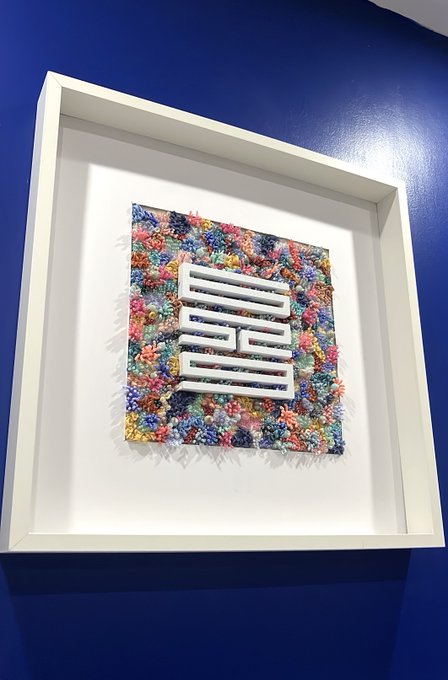 A framed artwork displayed on a blue wall, featuring a white stylized maze-like symbol or logo set against a textured, multicolored background of what appears to be plastic or shredded material.