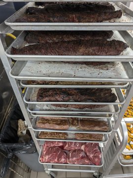 A metal rolling rack is stacked with sheet pans holding various cuts of raw and cooked meat, including ribs and steaks, in what appears to be a commercial kitchen setting.