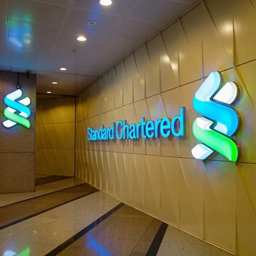 The image shows the illuminated "Standard Chartered" logo and name displayed on a gold-toned wall in what appears to be a corporate lobby or entrance area. This relates to the post by featuring Standard Chartered analyst Geoffrey Kendrick, who is commenting on potential partnerships between traditional finance and DeFi protocols like Uniswap and Robinhood.