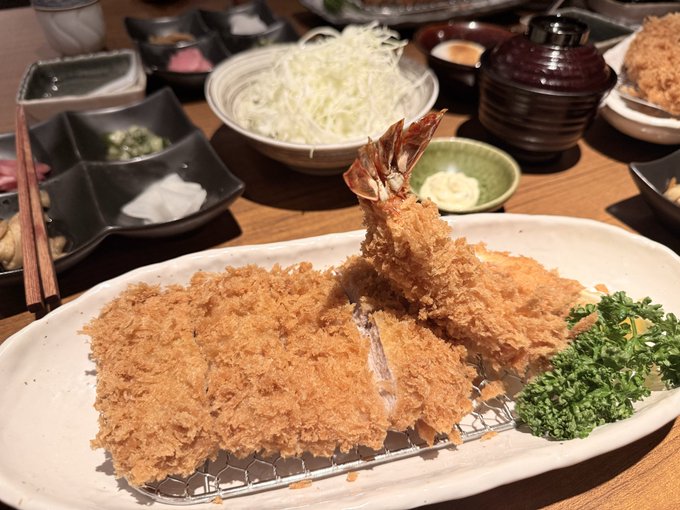 A Japanese tonkatsu meal featuring a breaded and fried pork cutlet with a large shrimp tail standing upright, served with shredded cabbage and various side dishes.