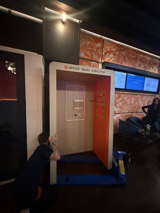 A photo of someone crouching and interacting with a small 'Office Hours Simulator' booth installation, which features an orange interior, in what appears to be a lounge or event space with monitors in the background.