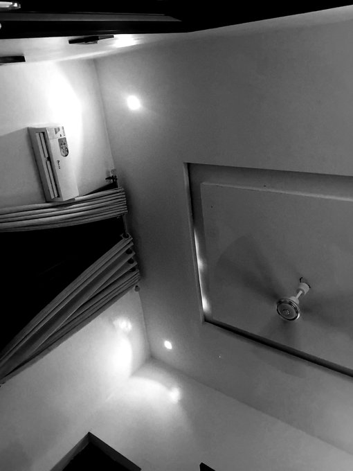 A black-and-white, sideways-oriented photograph of a bathroom corner showing an air conditioning unit, a showerhead, and recessed ceiling lighting.