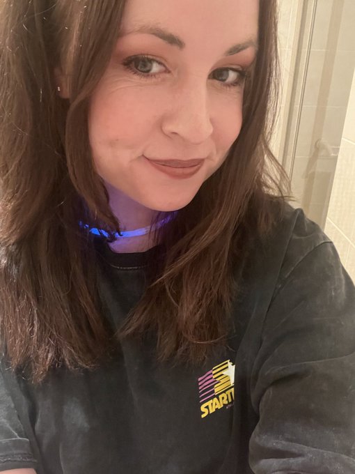 Close-up selfie of a smiling woman with long brown hair wearing a dark t-shirt featuring a pink and yellow 'STAR17' logo.