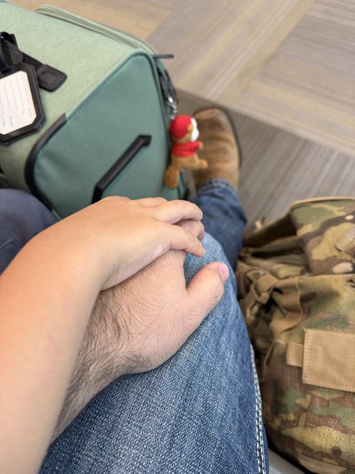 A first-person view looking down at a person's hands clasped on their lap wearing jeans, with a sage green suitcase, a small plush charm, tan boots, and a camouflage backpack visible nearby.