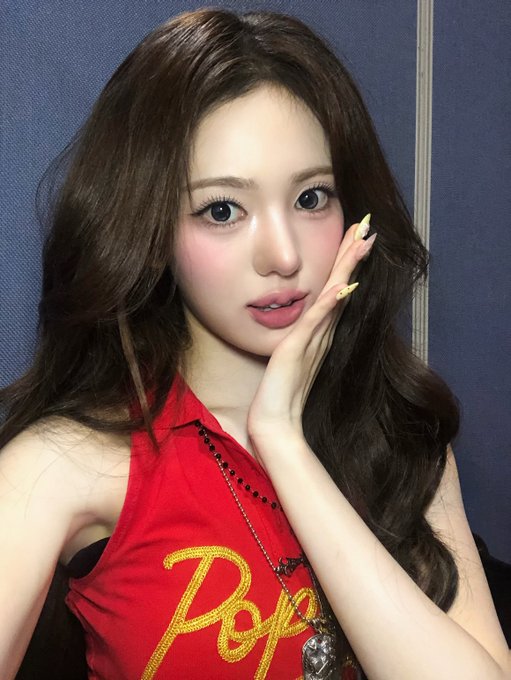 A young woman with long wavy hair poses with her hand near her cheek, wearing a red jersey with "Rayo" embroidered in yellow. The hashtags #KiiiKiii #키키 #SUI #수이 suggest she is Sui, a member of the K-pop group KiiiKiii, likely photographed during a casual backstage or promotional moment.